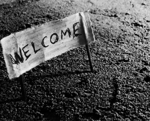 "Welcome" written on a piece of fabric, attached to a stick at either end with the sticks in the ground to create a sign