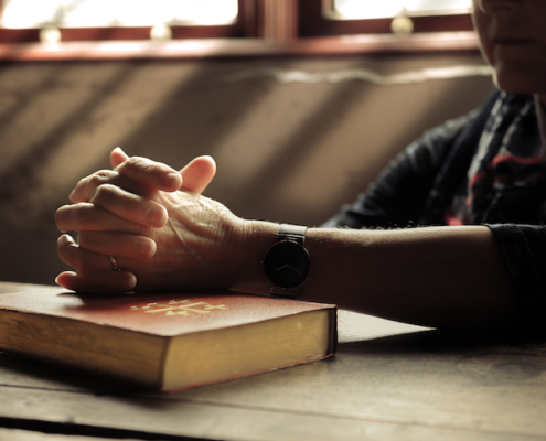hands folded as if in prayer sitting on top of a Bible with light streaming through a window in the background