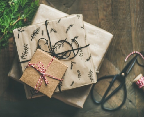 3 wrapped gift boxes stacked up from largest on the bottom to smallest on top all on a wooden table, with greenery on the left and scissors and twine on the right