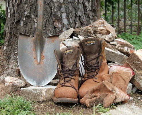 work boots, a pointed shovel and work gloves alongside a tree trunk and stone rubble