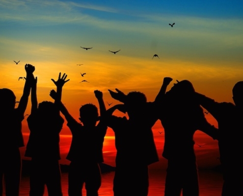 silhouettes of six children facing a sunset over water with hands raised and birds in the sky above